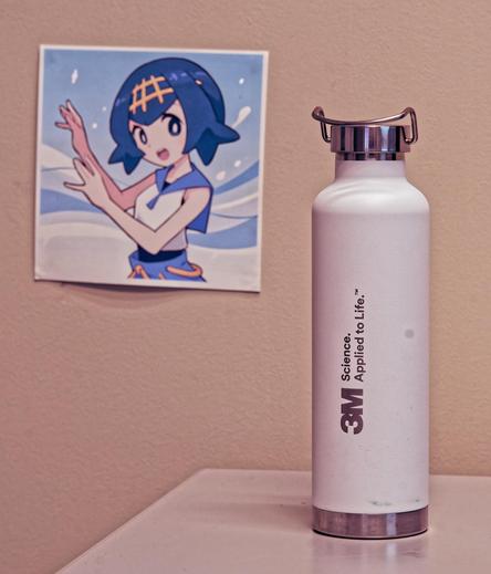 A white water bottle with chrome accents and a label that reads 3M: Science Applied to Life on a white table with a beige wall behind an a square image of an anime girl with blue hair and eyes dancing in front of a blue wave background