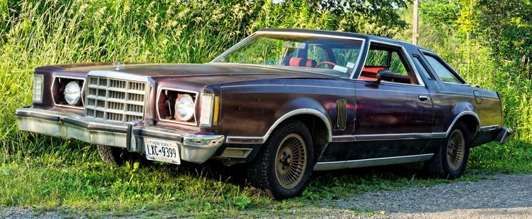 A maroon car with an absurdly long hood parked on the margin of grass and gravel with tall mature grass behind it and headlight doors stuck open;  I guess you're not supposed to post pictures of cars with license plates but...  it's my car