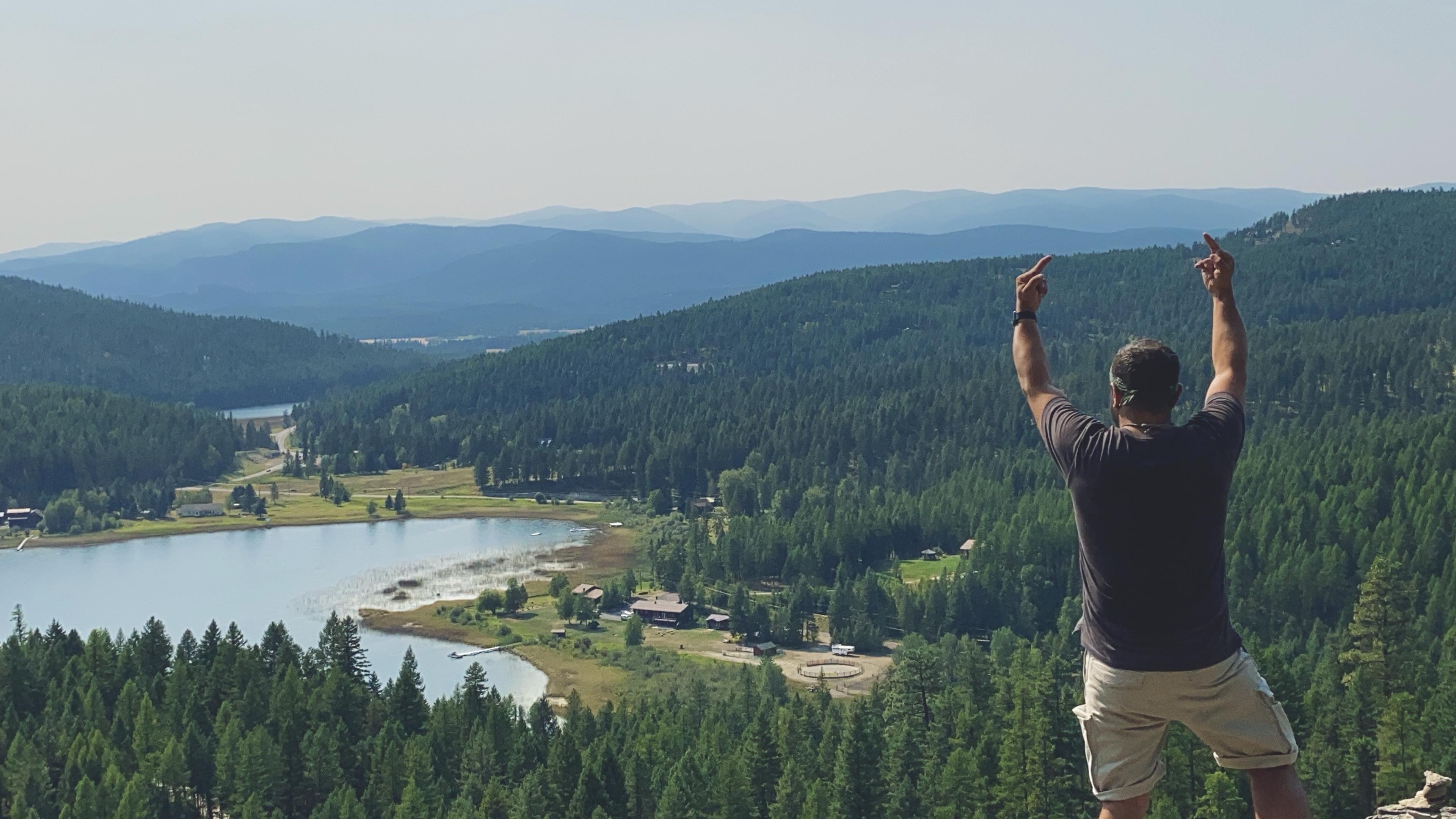 Me standing on a rocky outcrop with their back to the camera, raising both hands in a gesture of triumph (middle fingers). The background features a scenic view of a lush green valley with a lake, surrounded by forested mountains under a clear sky.