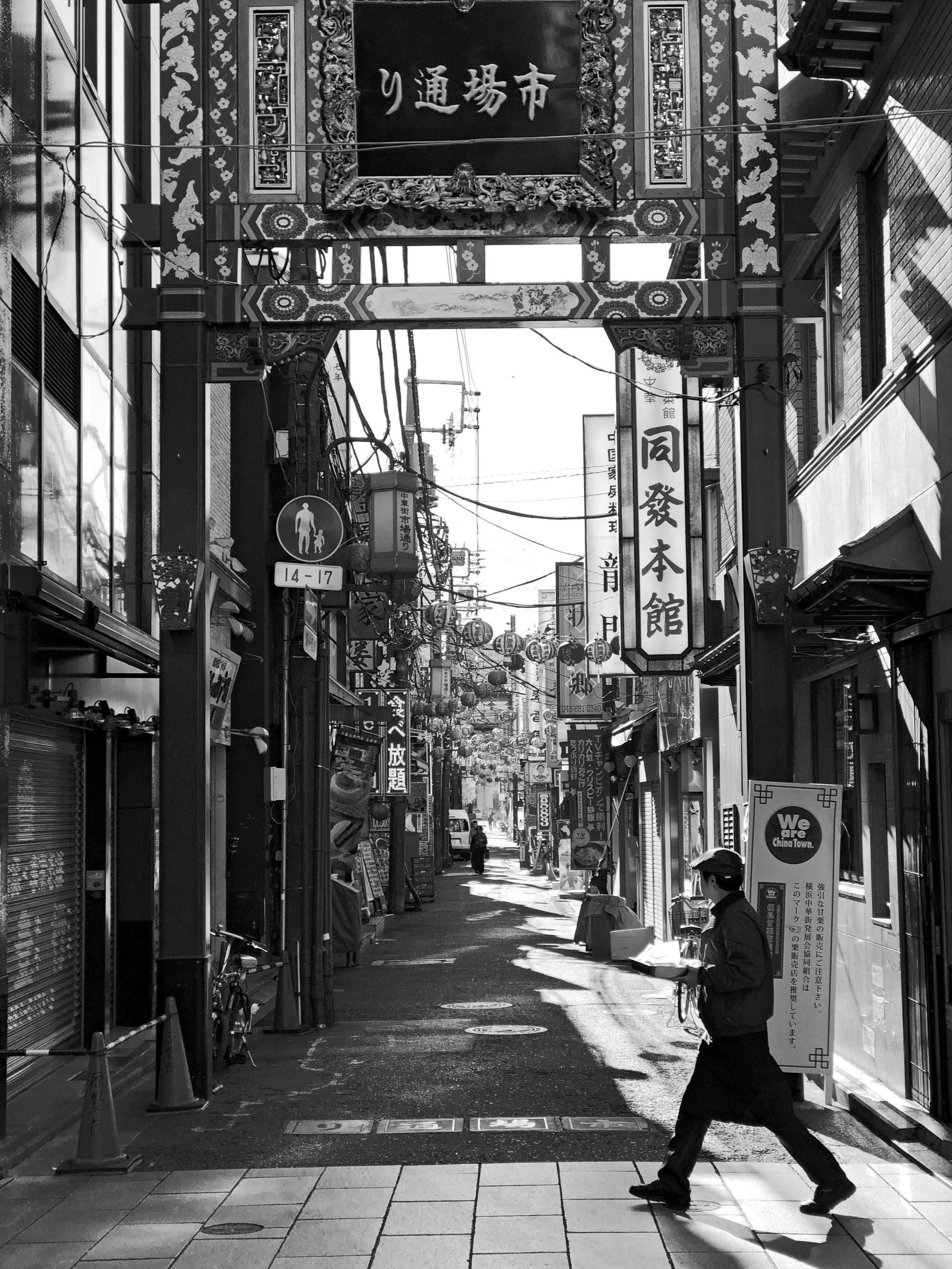 A black-and-white street scene in Chinatown featuring a decorative archway.  A person walks along a narrow alley lined with shops and signs in both Japanese and English. The street is partially illuminated by sunlight. 