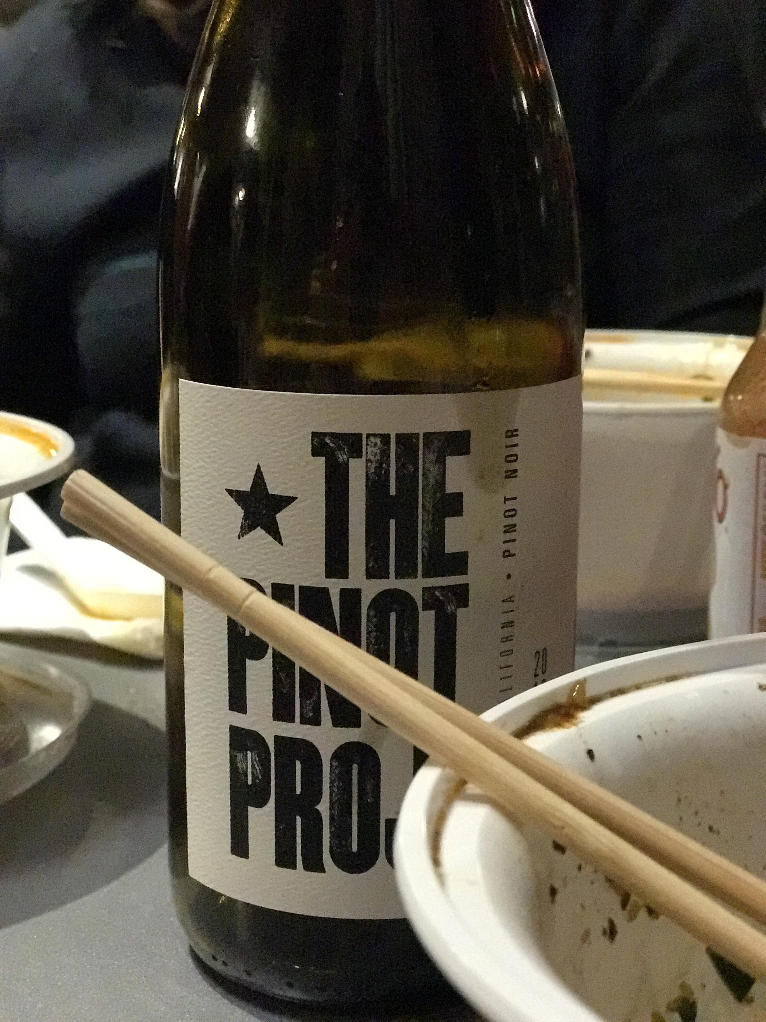 A bottle of wine labeled "THE PINOT PROJECT" next to a half-empty bowl with remnants of food and a pair of chopsticks resting on it.