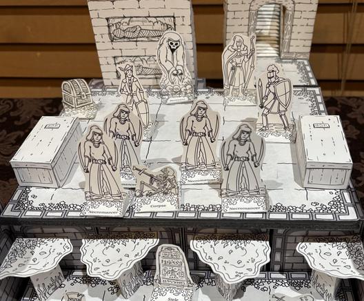 A papercraft scene from the game. Three heroes - an archer, a squire, and a warden - have entered a room with four necromancers and a ghostly revenant. In the foreground the floor drops away into a lower level filled with water. Around the room are a door, sarcophagi, burial niches, a corpse, and a treasure chest.