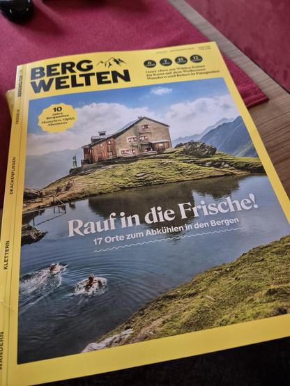 A yellow magazine is seen lying on a brown table, with a dominant color scheme of yellow and brown. The magazine appears to have a house on top of a hill on its cover. The setting seems to be outdoors, possibly near a lake, with water visible in the background.  The magazine is the main focus, showcasing a publication that may offer articles or stories related to outdoor living or nature.