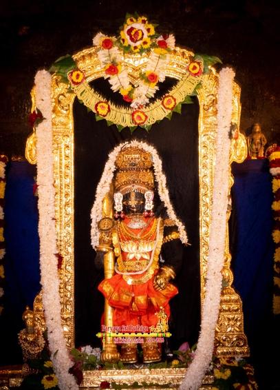 Idol of Hindu deity Krishna standing dressed in orange; wearing gold shoes, gold palm coverings, and several gold ornaments including a large necklace with a large lotus-shaped medallion. Holding a gold staff in right hand, left hand on left front waist. A string of white flowers shoulder-shoulder going over the crown. 

A gold arch around Krishna, extending quite high above the crown. A long string of white flowers along the sides of the arch making a U-shape. A string of areca flowers at the top of the arch along with a smaller string of white flowers.

Black background fills the arch. Dark blue cloth background behind the arch.