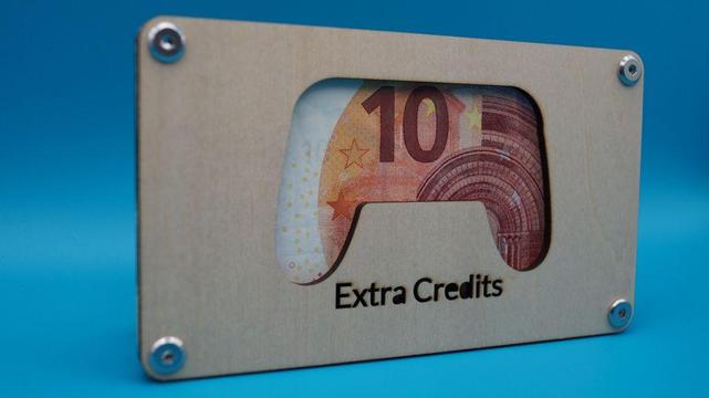 Zusammengebaute Holz-Geldgeschenkhalterung im Design eines Spielekontrollers mit der Gravur „Extra Credits“. Durch das Sichtfenster ist ein gefalteter 10-Euro-Schein sichtbar.