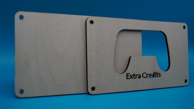 Zwei gelaserte Holzplatten im Stil einer Spielekonsole, die durch ein ausgeschnittenes Sichtfenster das gravierte Wort „Extra Credits“ und die Form eines Kontrollers zeigen. Die Platten sind noch nicht montiert.