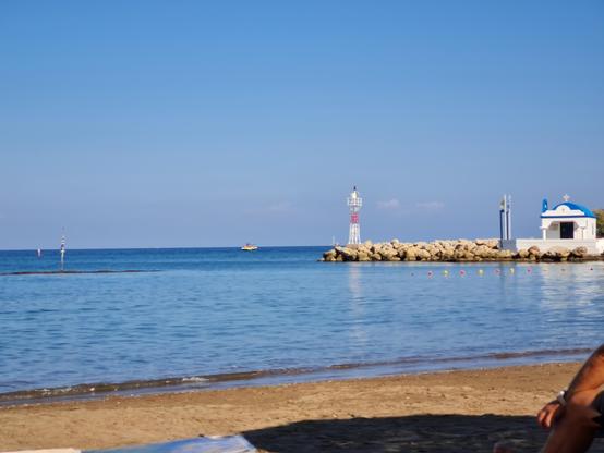 Es ist ein blauer Himmel und das Meer sowie etwas vom Strand zu sehen. Auf der rechten Seite gibt es zwischen Wasser und Himmel etwas Land mit einem Leuchtturm und einer kleinen Kapelle.
