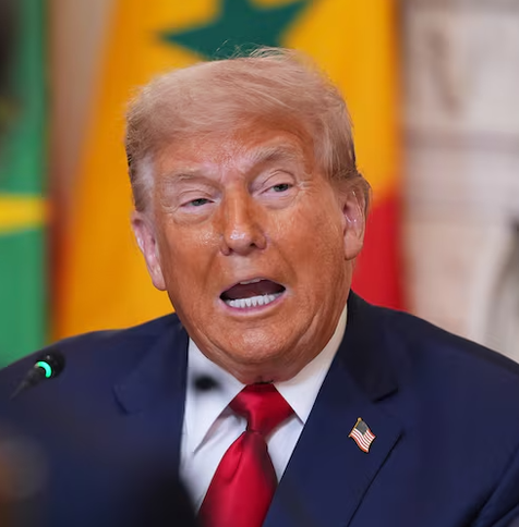The image features a Trump with light-colored hair, wearing a dark blue suit, a white shirt, and a red tie. He is speaking into a microphone, with his mouth open, suggesting he is in the midst of delivering a speech or statement. On the lapel of his suit jacket, there is a small American flag pin. The background is blurred, but it appears to include a flag with green, yellow, and red colors, possibly indicating a formal or governmental setting. The man's expression is serious, his face is orang…