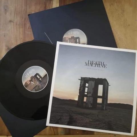 Eine Schallplatte mit dem Titel "Death Machine" liegt auf einer hölzernen Unterlage, begleitet von ihrer Hülle. Das Cover zeigt eine trostlose Landschaft mit einem zerfallenden Gebäude vor dem Hintergrund eines Sonnenuntergangs. Eine zweite Hülle mit demselben Design ist leicht sichtbar.