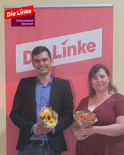Zwei Personen stehen vor einem Die-Linke-Rollup und halten Blumensträuße. Links: Jannick Metz, Oberbürgermeisterkandidat für Bayreuth. Rechts: Juliane Glaschke, Landratskandidatin für den Landkreis Bayreuth. Beide lächeln in die Kamera.

