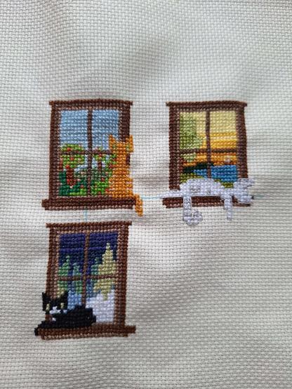 Picture of a work of cross stitch, it's a small and stylistic piece without a lot of shading. The fabric is an off white color. There are three windows depicted with cats sitting in front of them. Upper left is an orange cat and the outdoors is trees with blossoms. Upper right is a white and gray cat with a  yellow sky, a tree and a small boat. Down left is a black cat in front of a window with snow and fern trees in the background. 