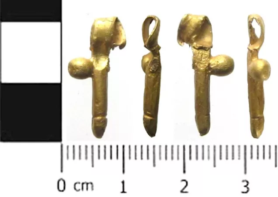 Photograph of a tiny golden phallus pendant against a measuring scale. It's about 2 cm in height and 2 mm in width.