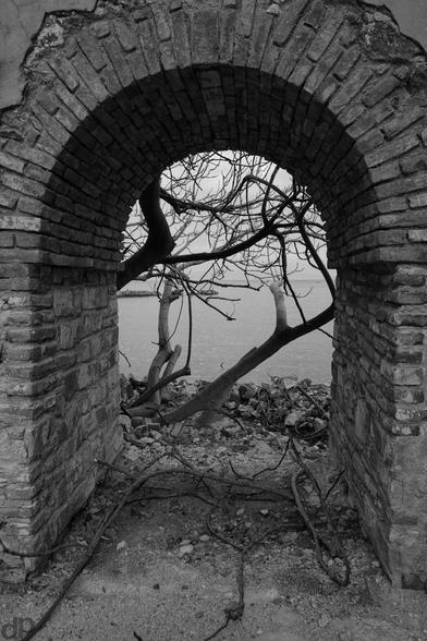 Schwarz-Weiß- Fotografie: 
Eine aus Ziegelsteinen gebaute Tür mit Bogen, darum herum abgebrökelter Putz. Es gibt kein Türblatt und der Blick fällt auf einige kahle Äste und dahinter auf das Meer.
Auf dem Boden liegen alte Äste und Steine.