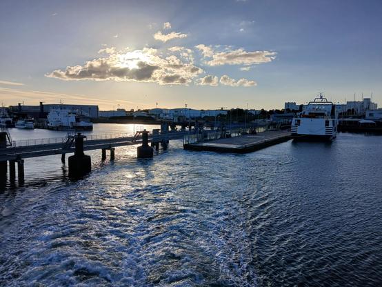Départ du port de Lorient dans la lumière de la fin d'après-midi. Soleil derrière les nuages, un ferry à quai. Photo prise depuis le ferry partant.