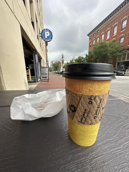 Color still life photo of to go coffee cup and a crumpled up paper wrapper on the corner of a picnic bench. A brick building is visible in the background to the right, a yellow building to the left has a circular “P” parking sign on it. Between is a cloudy, threatening sky.