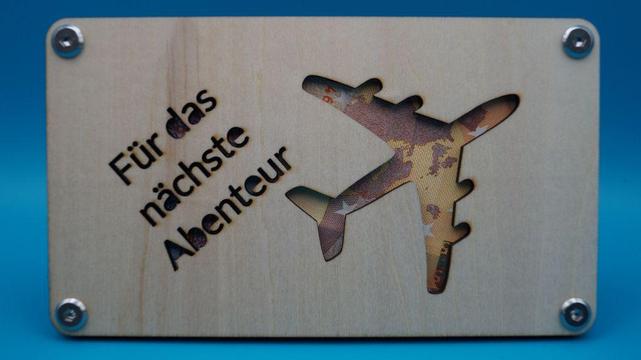 Holzrahmen mit Flugzeug-Ausschnitt und dem gelaserten Schriftzug „Für das nächste Abenteuer“, dahinter ein 10-Euro-Schein sichtbar eingesetzt