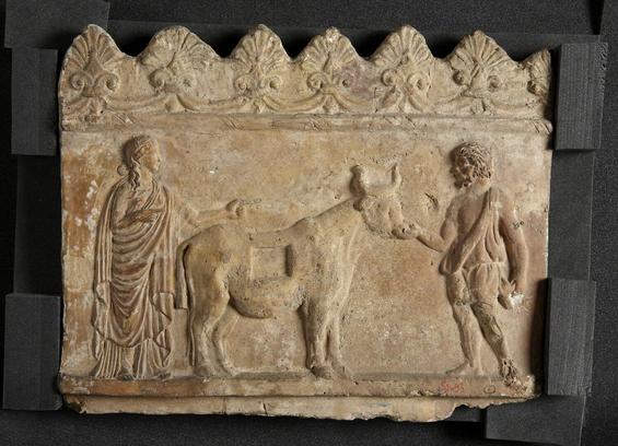 Relief with palmettes on top, depicting Daedalus and Pasiphae. Pasiphae wears a chiton and a himation. She is standing at the rear of the fake cow created by Daedalus. Daedalos touches the cow by the muzzle. He is bearded and wears an exomis.