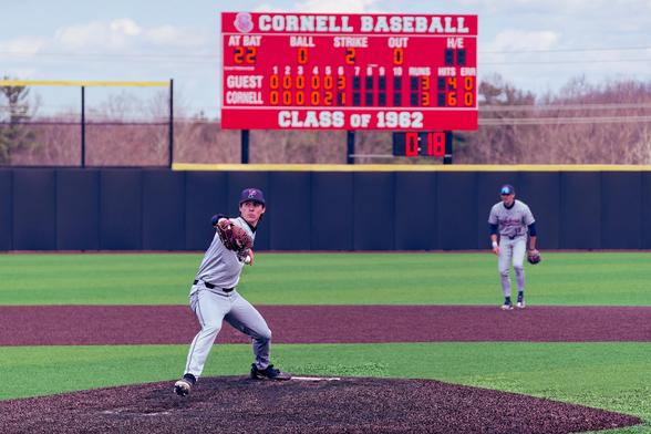 Baseball pitcher winds up with light colord jersey and shorts and a blue cap with an outfield in the backgrround and a scoreboard that says CORNELL BASEBALL in the sixth inning with 2 strikes.