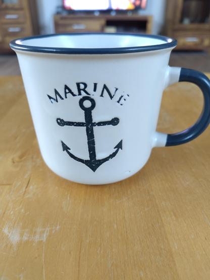 Das Bild zeigt eine Tasse mit einem maritimen Design. Die Tasse ist hauptsächlich weiß mit einem dunkelblauen Rand und einem dunkelblauen Griff. Auf der Vorderseite der Tasse ist ein schwarzes Anker-Design abgebildet, über dem das Wort "MARINE" in Großbuchstaben steht. Die Tasse steht auf einer hellbraunen Holzoberfläche.