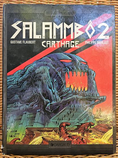 The cover of Philippe Druillet's Salammbô 2 - Carthage, a Lone Sloane story published in 1982, inspired by Gustave Flaubert's book Salammbô. Cover art features a monstrous monument figuring some evil deity, with red eyes and mouth where a fire burns. Tiny human figures seem to be praying below it. Two little reptilian monsters swim in the foreground. A city is seen in the background. Published by Dargaud in their Histoires Fantastiques collection.