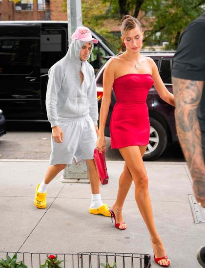 Justin und Hailey Bieber. Sie farblich abgestimmt in einem schicken roten Kleid mit passenden Schuhen, er in einer Art kurzhosigen Jogginganzug, auf der Kapuze eine rosa Cap, gelbe Crocs tragend.