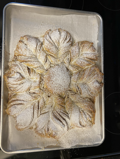 Viewer is looking down on a baking sheet from above. The aluminum baking sheet is lined with parchment paper and sits on a dark glass range top. The star bread has eight puffy points or petals surrounding a round center. Each point is formed of two twists of dough layered cinnamon sugar and pinched together at the tip. The whole thing is golden brown and sprinkled with confectioner’s sugar.
