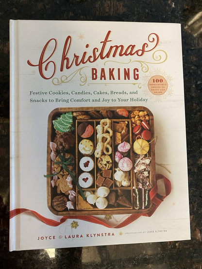 Cover of the cookbook. The background is white. Main title in red is Christmas Baking. Subtitle in green is Festive Cookies, Candies, Cakes, Breads, and Snacks to Bring Comfort and Joy to Your Holiday. A square wooden tray holds a variety of Christmas goodies. Joyce and Laura Klynstra are the authors. The book is on a dark granite counter which reflects some light.