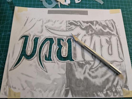 Exacto knife being used to cut out thai letters saying Muay Thai from a sheet of paper.