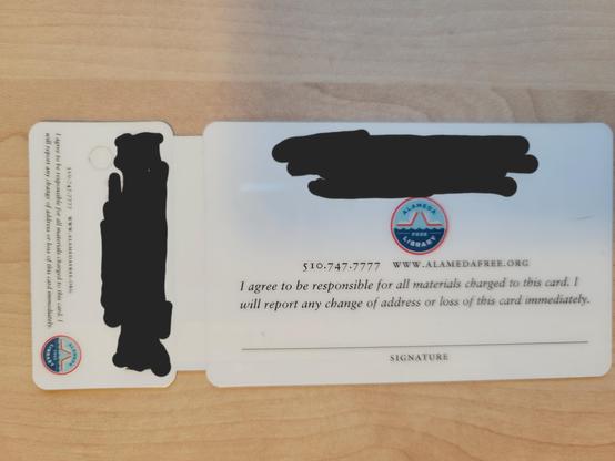 Reverse of both library cards. At center of the larger card is the circular logo of the Alameda Free Library: the bottom is water, while two partially-open books in the top half seem to form a raised drawbridge under a light-blue sky. The name of the library appears in the background: "Alameda" (top, blue) and "Free Library" (bottom, white). The bar codes and library card number are blacked out. The bottom of the card has contact details and a blank signature line under an "I agree to be responsible...." declaration.