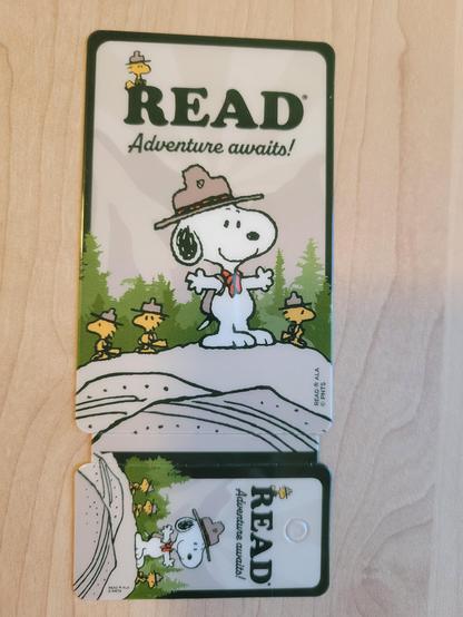 Front of 2-part library card (credit-card-sized attached to keychain-sized card). Both show Snoopy dressed as a Beagle Scout with distinctive brown peaked hat, brown backpack, and red neck kerchief. Four yellow Woodstock birds are dressed in similar fashion. Snoopy and 3 birds are standing outdoors on a what might be a group of boulders. In the background are the tops of fir trees, with mountains rising high above them to each side. A 4th bird sits at top on the text "Read" (bold serif block letters). Text "Adventure awaits!" follows in small sans-serif script. Both cards have the same design.
