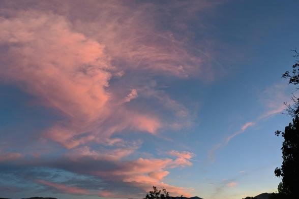 A photo of the sky after sunset, there are some pink clouds along with the darkening blue sky. It is composed of 3 portrait images combined as a panorama. There are some bits of trees, mountains, hills on the bottom and right sides.