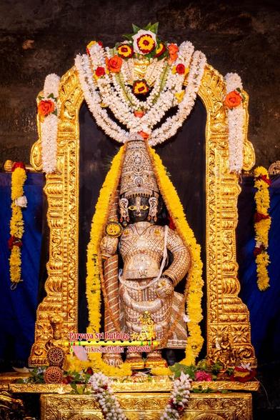 Idol of Hindu deity Krishna a diamond-clad Krishna standing with a matching crown and a similarly clad staff in right hand. Krishna stands on an ornate gold box and is surrounded by a gold arch. A string of yellow flowers around Krishna. Strings of white flowers hanging down at the top of the arch. A string of white flowers in U shape on the box at Krishna's feet. Blue cloth background with strings of yellow flowers hanging down at both ends.