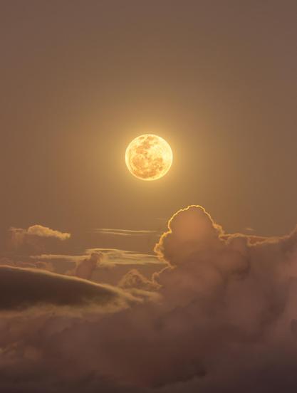 A golden Full Moon rises behind layers of drifting clouds, casting warm light across the sky. The image, taken from Bahia, Brazil, combines two exposures to reveal both fine details on the Moon's surface and subtle rainbow-like iridescence in the cloud edges. The sky is darkening, creating a dramatic contrast between the Moon and the silhouetted clouds.