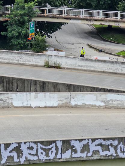 Vertical color photograph of a several empty highway overpasses viewed from slightly above so that you can see the road surfaces. Near the center of the image, a lone person walking is visible in the distance, wearing a hi-viz yellow vest. Towards the left is a small highway sign with “83 SOUTH” and “25 MPH” on it. The sign is surrounded by a rough growth of small trees. At the top of the image, a foot bridge traverses horizontally across the frame. At the bottom, a Jersey barrier features a scrawl of white painted graffiti.