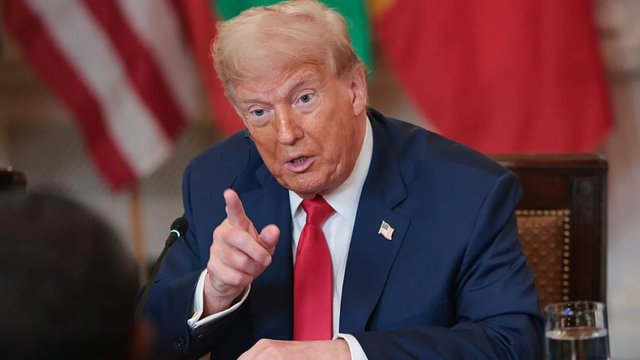 The image shows a Tariff man in a formal setting, likely a meeting or press conference. He is wearing a dark blue suit with a white shirt and a red tie. A small American flag pin is visible on his left lapel. The man is gesturing with his right hand, pointing with his index finger, suggesting he is making a point or emphasizing a statement. The background includes a blurred American flag and another flag with red and green colors, indicating a formal or governmental context. A glass of water is…