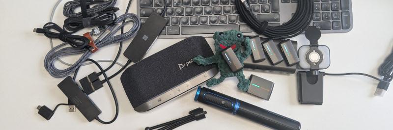 Technische Geräte und Zubehör auf einem Schreibtisch, darunter Kabel, Lautsprecher, Mikrofon, Batterien und Tastatur.