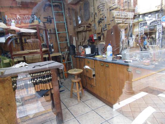 Blick durch das Schaufenster eines Instrumentenbauers. Teilweise sind die zu sehenden Werkzeuge ordentlich aufgehängt, aber auf dem Tresen liegen diverse Utensilien durcheinander.