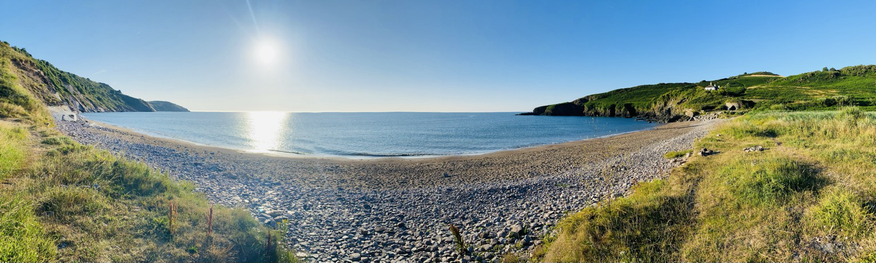 A panoramic photo of Mansands beach looking out towards the horizon, the sun still rising in the sky. The beach is deserted. Pebbles in the foreground then sand and sea