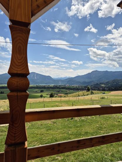 A serene scene captured in this image showcases a wooden post standing tall in the foreground, with a vast field stretching out behind it. In the distance, majestic mountains rise up against the clear blue sky, creating a stunning backdrop. The wooden post adds a rustic charm to the landscape, blending seamlessly with the natural surroundings. The field is lush with green grass, and fluffy clouds float peacefully overhead. This tranquil outdoor setting is perfect for those seeking solace and co…