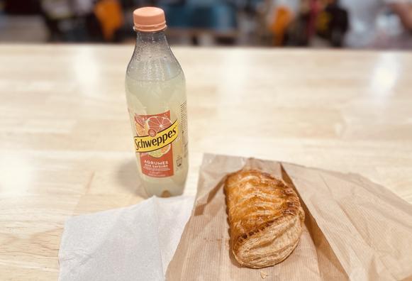 Ein PET-Fläschchen Schweppes «Agrumes aux Saveurs», daneben liegt eine halbmondförmige Apfeltasche mit üppiger Glasur.
