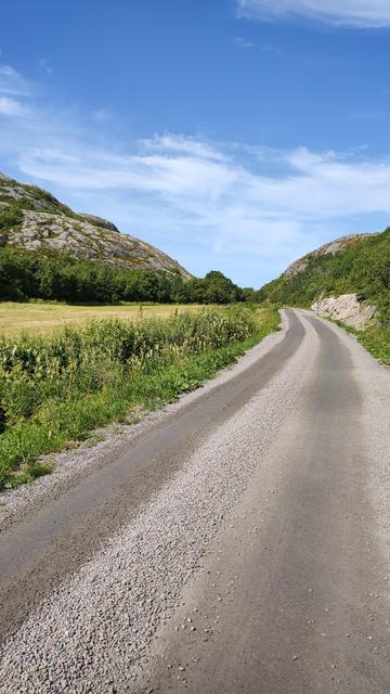 Unbefestigte Straße in Norwegen, blauer Himmel, links ein grünes Feld und felsige Hügel.