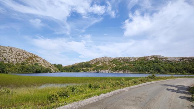Eine unbefestigte Straße in Norwegen, dahinter grüne Moorlandschaft, ein See, felsige Hügel und blauer Himmel mit ein paar Wolken.