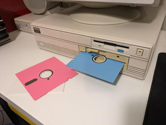A Tandy desktop computer with a pastel blue 5.25" floppy disk sticking out, and a pink disk sitting in front of it.