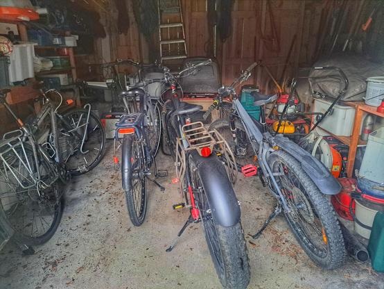 Kuva vajan sisältä. Etualalla neljä polkupyörää (cyckocross, kaupunkisähköpyörä, såhköläski, maastopyörö). Näiden takana ajettava ruohonleikkuri, pari työnnettävää ruohinleikkuria, oksasilppuri ja muita puutarhakoneita.