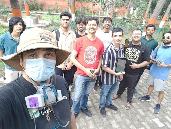 A selfie taken by a person wearing a hat, a surgical mask, and with a elastomeric respirator hanging around their neck.

Behind them are 9 boys looking at the camera, some holding up their phones and tablets to show different OSM applications.

All are standing in a park.