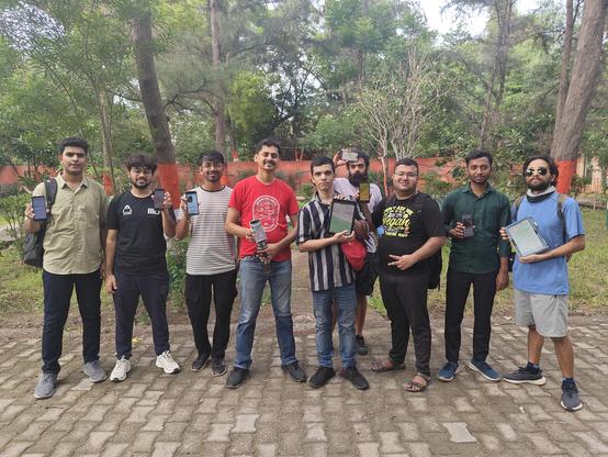 9 boys standing on a park path, smiling and facing the camera. They are holding up their phones and tablets to show applications like MapComplete and CoMaps.