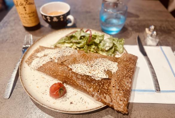 Eine Galette, salziger, dreieckig umgeschlagener Buchweizen-Pfannkuchen. Dazu etwas Salat und eine kleine gebratene Cherry-Tomate. Dahinter steht eine Tasse mit bretonischem Apfelwein (Cidre).