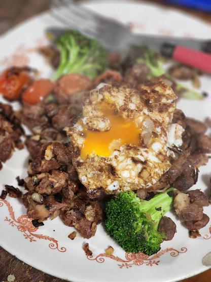 A fried egg, very yellow yolk broken, on a pile of brown beans, with fried cherry tomatoes and broccoloi florets on the side. 