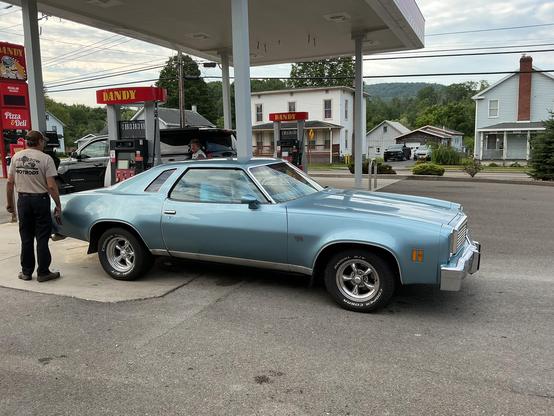 A man facing away from the camera stands near the back of a blue two door car on a concrete pad by a filling station.
