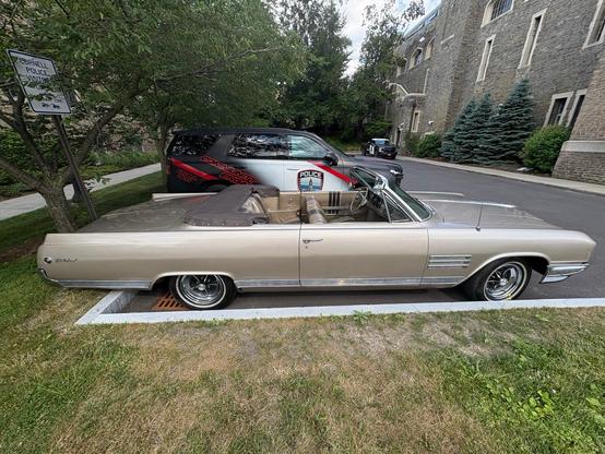 Beige convertible car with finned style on a long trunk and long hood at the near corner of a parking lot with worn grass in front, a police SUV in back and a row of trees and a building with a stone side